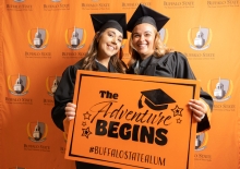 Two students in cap and gown holding sign that reads "The Adventure Begins"